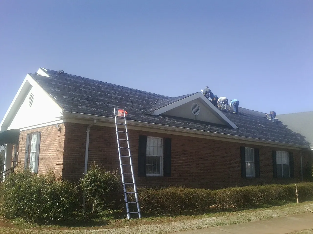 Crew tearing shingles off a brick ranch roof with a ladder set — Emergency Roof Repair work by our crew in Deckerville